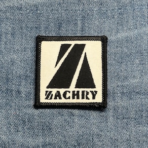 May include: Square patch with a white background and black geometric design. The word "ZACHRY" is printed in black, block letters below the design. The patch has a black border and is set against a blue denim background.