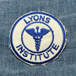 May include: Round patch with the words "LYONS INSTITUTE" in blue lettering. A blue caduceus symbol is in the center. The patch has a white background with a blue border, set against a denim background.