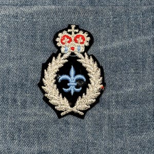May include: Embroidered patch featuring a crown with red and blue accents atop a black background. A silver wreath surrounds a blue fleur-de-lis. The patch is set against a blue denim background.