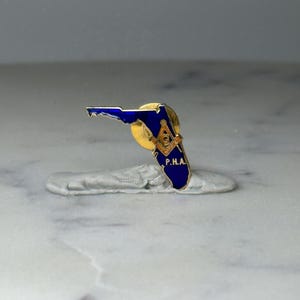 May include: A blue and gold enamel pin in the shape of the state of Florida. The pin features the letters "P.H.A." and a Masonic symbol. The pin is set on a gold-colored metal base.