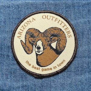 May include: Circular patch with a brown ram's head and the text "ARIZONA OUTFITTERS" and "the best game in town" in a brown border. The patch is on a blue denim background.