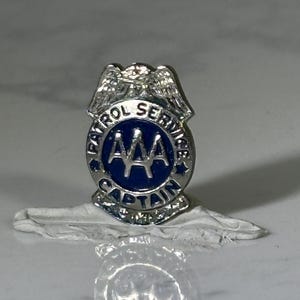 May include: A silver-toned AAA Patrol Service Captain pin. The pin features a blue circular center with the AAA logo and the words "PATROL SERVICE" and "CAPTAIN" in a circular pattern. The pin has a pair of wings at the top.