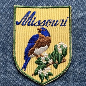 May include: Embroidered patch featuring a bluebird perched on a flowering branch, set against a yellow background. The word "Missouri" is embroidered in blue script above the bird. The patch has a green border and is set on a blue denim background.