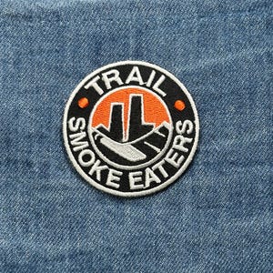 May include: A circular patch with the words "TRAIL SMOKE EATERS" in a white, bold font. The patch features an orange and black graphic of a mountain scene. The patch is sewn onto a blue denim fabric.