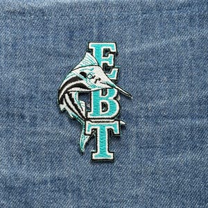 May include: Embroidered patch featuring a marlin fish design in shades of teal, black, and white. The patch includes the letters "EBT" in a teal and white outline font. The patch is set against a blue denim background.