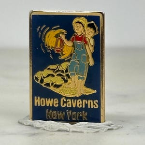 May include: A rectangular Howe Caverns New York pin with a blue background and gold trim. The pin features two children, one carrying a lantern. The text "Howe Caverns New York" is in gold.