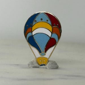 May include: A colorful hot air balloon pin with a gold border. The balloon features sections of blue, red, yellow, and white. The pin is set against a neutral background.