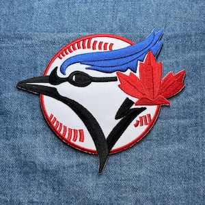 May include: Embroidered patch featuring a stylized blue jay head with a black beak and eye, blue feathers, and a red maple leaf. The design is set against a white baseball with red stitching and a red border. The patch is on a denim background.