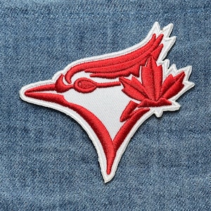 May include: Embroidered patch featuring the Toronto Blue Jays logo. The patch has a white background with a red bird head and a red maple leaf. The patch is on a blue denim background.
