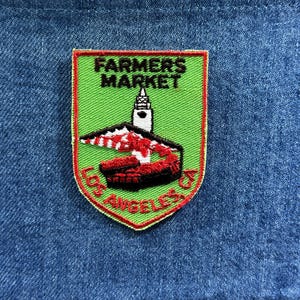 May include: Embroidered patch with a green background and red border. The patch features the text "FARMERS MARKET" above a graphic of a building and a red and black structure. The text "LOS ANGELES, CA" is arched at the bottom.