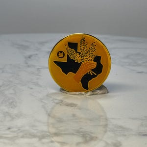 May include: A round, gold-colored pin featuring a black silhouette of the state of Texas. A hand holds a bundle of wheat, with a small logo in the upper left. The pin is set against a light background.