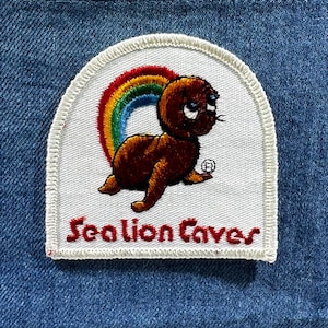 May include: Embroidered patch featuring a brown cartoon seal with a rainbow and the text "Sea Lion Cove" in red. The patch has a white border and is set against a blue denim background.