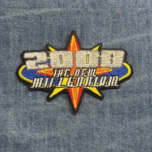 May include: Embroidered patch with the text "2000 The New Millennium" in a stylized design. The patch features a starburst shape with red, orange, and yellow colors, and the text is in blue and silver. The patch is on a denim background.