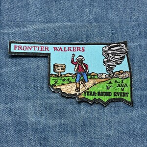 May include: Embroidered patch in the shape of Oklahoma with a cowboy figure, a tornado, and the text "FRONTIER WALKERS" and "YEAR ROUND EVENT". The patch features a blue sky, green grass, and a brown road.