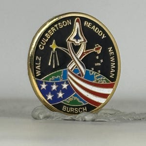 May include: A round, gold-rimmed pin with a black background featuring a stylized rocket, stars, and the American flag. The pin has the names WALZ, CULBERTSON, READDY, NEWMAN, and BURSCH around the edge.