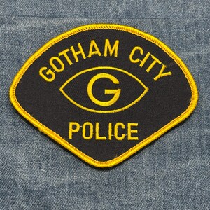 May include: A black and yellow Gotham City Police patch. The patch has a yellow border and features the words "Gotham City Police" in yellow, along with a yellow eye design with a "G" in the center. The patch is diamond-shaped.