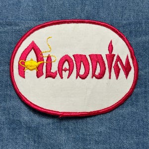 May include: Embroidered patch featuring the word "Aladdin" in pink, with a yellow genie lamp. The patch has a white oval center with a pink border, set against a blue denim background. A decorative item.