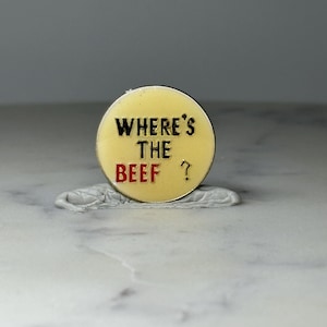 May include: A round, yellow enamel pin with the phrase "WHERE'S THE BEEF?" in black and red lettering. The pin has a silver-colored backing and is set against a neutral background.