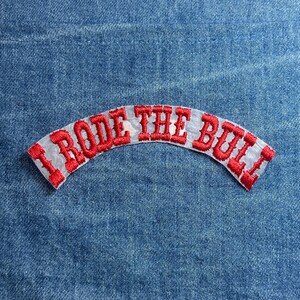 May include: Embroidered patch with the text "I Rode The Bull" in red letters on a white background. The patch is arched and set against a blue denim fabric.