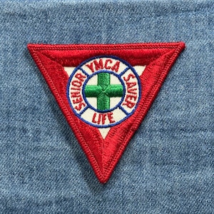 May include: A red, triangular patch with white stitching and a blue and white circular emblem. The emblem features a green cross and the words "YMCA SAVER LIFE". The patch is set against a blue denim background.