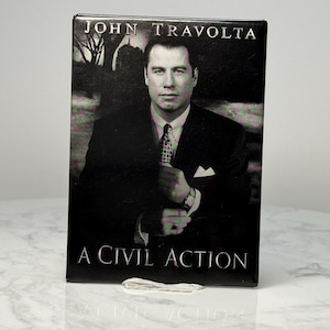 May include: A rectangular black DVD case featuring John Travolta. The cover art includes a portrait of Travolta in a suit, with the title "A Civil Action" in white text. The top of the case reads "John Travolta".