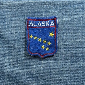 May include: A blue and red Alaska patch with the word "ALASKA" in white lettering. The patch features a blue background with yellow stars, and a red border. The patch is on a blue denim background.