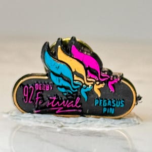 May include: A vintage Pegasus pin from the '92 Derby Festival. The pin features a black oval shape with gold trim, and colorful wings in teal, yellow, and pink. Text includes "92 Derby Festival" and "Pegasus Pin."