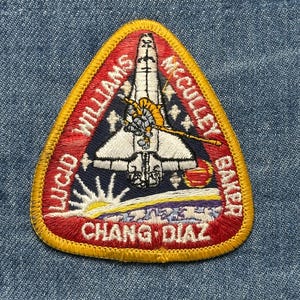 May include: Embroidered patch with a triangular shape, featuring a space shuttle design. The patch has a red border with gold trim and includes the names WILLIAMS, MCCULLEY, BAKER, CHANG DIAZ, and LUCID. The background is navy blue.