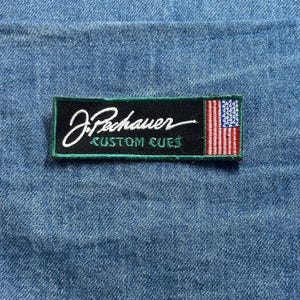 May include: A rectangular patch with the text "J. Pechauer CUSTOM CUES" in white script and block letters on a black background, framed by a green border. The patch includes a small American flag on the right side. The patch is set against a blue denim background.