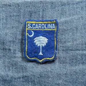 May include: A blue and gold embroidered patch featuring the state of South Carolina's symbols. The patch has the text "S.CAROLINA" at the top, a crescent moon, and a palmetto tree.