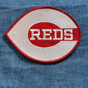 May include: Embroidered patch featuring the Cincinnati Reds logo. The patch has a white background with a red oval and the word "REDS" in white block letters. The patch is outlined in red and is set against a blue denim background.