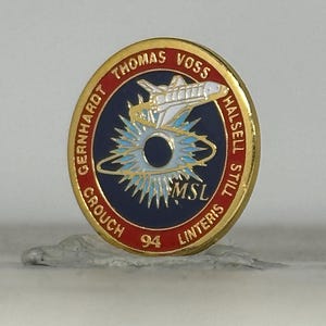 May include: A gold-colored pin with a red border featuring the names Thomas Voss, Gernhardt, Crouch, Linters, Halsell, and Still. The pin's center has a blue background with a white and yellow space shuttle and a stylized sunburst design. The text "MSL" and the number "94" are also visible.