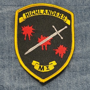 May include: A black shield-shaped patch with a yellow border and text that reads "HIGHLANDERS" and "MI". The patch features a silver sword and three red bursts.