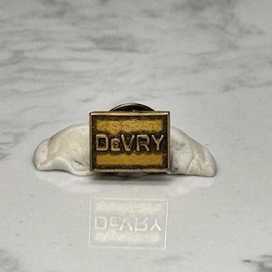 May include: A gold-toned pin with the word "DeVRY" in bold, black letters on a rectangular background. The pin is set against a white, textured surface with a marble-like pattern.
