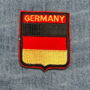 May include: Embroidered patch featuring the German flag design. The patch has a shield shape with a black border. The top section is red with the word "GERMANY" in gold lettering. Below are black, red, and gold horizontal stripes.