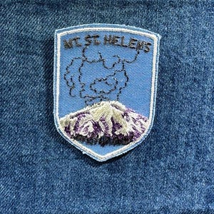 May include: Embroidered patch featuring Mt. St. Helens. The patch has a light blue background with a white border. The mountain is depicted with purple and white embroidery, and the text "MT ST HELENS" is stitched in silver.