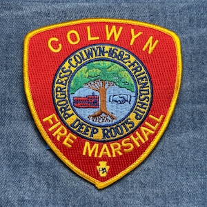 May include: A red and yellow embroidered patch with the text "COLWYN FIRE MARSHALL" in yellow. The patch features a circular emblem with a tree, handshake, and the words "PROGRESS-COLWYN-1682-FRIENDSHIP DEEP ROOTS".