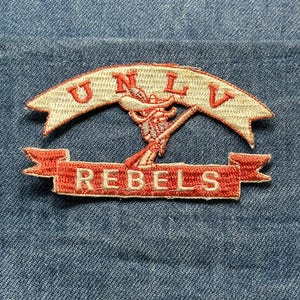 May include: Embroidered patch featuring the text "UNLV" in red letters on a cream-colored banner, and "REBELS" in red on a separate banner. A red and cream-colored mascot is in the center. The patch is on a blue denim background.