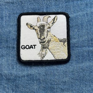 May include: Embroidered patch featuring a detailed goat illustration in shades of gray and tan, set against a white background. The word "GOAT" is printed in black below the goat. The patch has a black border.