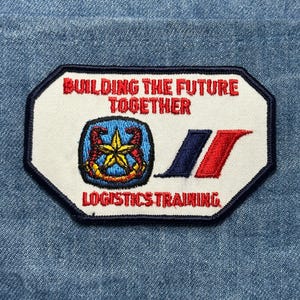 May include: A rectangular patch with a white background and a navy blue border. The patch features the text "BUILDING THE FUTURE TOGETHER" in red, and "LOGISTICS TRAINING" in red. A blue and gold emblem is on the left, with blue and red stripes on the right.