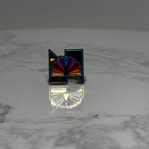 May include: A small, colorful pin featuring a stylized design with a rainbow fan shape and a geometric border. The pin has a silver-colored metal backing and is set against a neutral background.