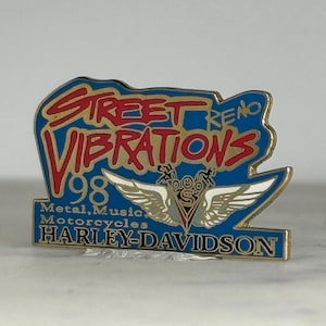 May include: A rectangular pin with a blue background and gold trim. The pin features the words "Street Vibrations '98" in red and yellow, along with "Metal, Music, Motorcycles" and "Harley-Davidson". A silver winged emblem is also present.