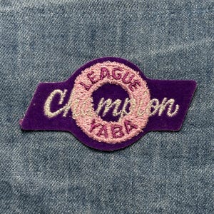 May include: A purple patch with the words "LEAGUE Champion YABA" embroidered in white and pink. The patch has a rectangular shape with a circular design in the center. The patch is on a blue denim background.