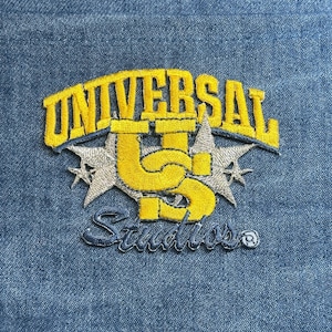 May include: Embroidered patch featuring the text "UNIVERSAL Studios" in yellow and silver. The design includes the letters "US" in yellow, with silver stars on a blue denim background. The patch is likely for apparel.