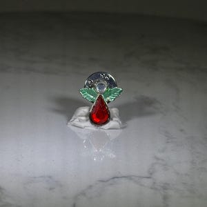 May include: A silver-toned brooch with a red teardrop-shaped gemstone. The brooch features a circular design with a clear stone and green leaf accents. The brooch is set on a white base.