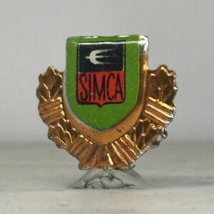 May include: A vintage SIMCA emblem pin. The pin features a shield design with a green background, a black and white emblem, and the word "SIMCA" in red. The shield is surrounded by a gold-colored wreath.