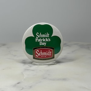 May include: A round white button with a green shamrock design and the text "Schmidt Patrick's Day." The Schmidt Beer logo is at the bottom. The button is on a white surface.