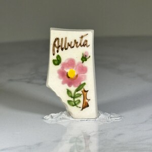 May include: A small, rectangular ceramic piece with the word "Alberta" in brown script. A pink flower with a yellow center and green leaves is painted on the front. The piece has a broken edge.