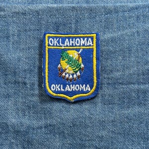 May include: A blue and yellow embroidered patch featuring the word "OKLAHOMA" at the top and bottom. The patch has a central design with a yellow circle, green leaves, and brown feathers.