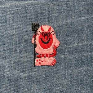 May include: Embroidered patch featuring a cartoon devil with red skin, horns, and a smiling face. The devil is holding a black pitchfork. The patch is on a blue denim background.